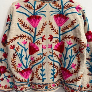 May include: A beige long-sleeve jacket featuring vibrant floral embroidery in shades of hot pink, deep red, and bright blue. The design includes symmetrical flower motifs, leafy stems, and a decorative border along the hem and collar area.