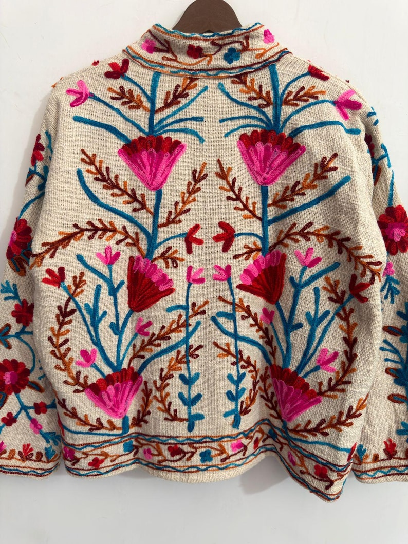 May include: A beige long-sleeve jacket featuring vibrant floral embroidery in shades of hot pink, deep red, and bright blue. The design includes symmetrical flower motifs, leafy stems, and a decorative border along the hem and collar area.