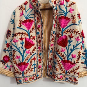May include: A cream-colored jacket featuring vibrant floral embroidery in shades of hot pink, deep red, and bright blue. The garment has a stand-up collar, long sleeves with folded cuffs, and an interior lining with a yellow and white vertical stripe pattern.