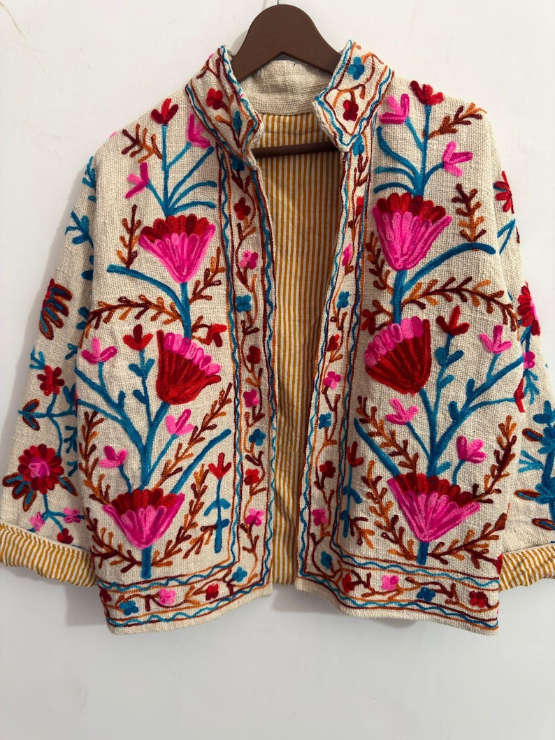 May include: A cream-colored jacket featuring vibrant floral embroidery in shades of hot pink, deep red, and bright blue. The garment has a stand-up collar, long sleeves with folded cuffs, and an interior lining with a yellow and white vertical stripe pattern.