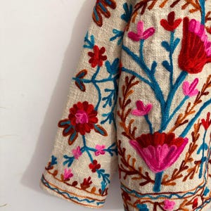 May include: A beige jacket featuring vibrant floral embroidery in shades of red, pink, orange, and teal. The design includes intricate botanical patterns with stems, leaves, and blossoms covering the fabric, finished with a decorative border.