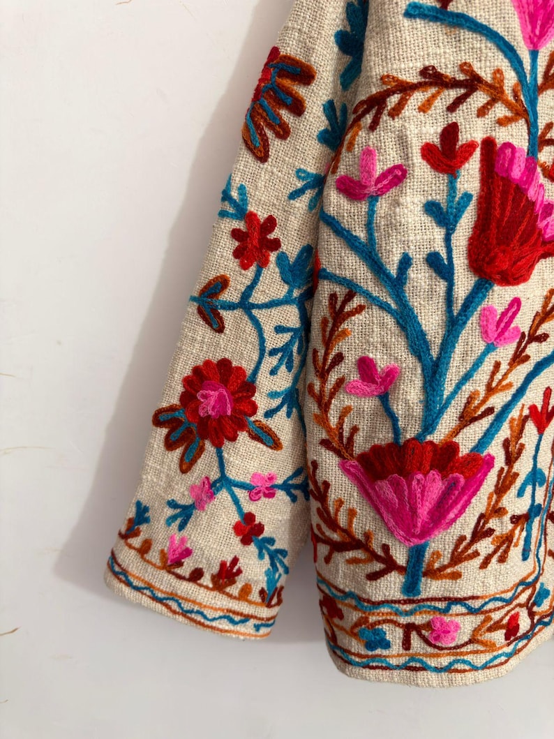 May include: A beige jacket featuring vibrant floral embroidery in shades of red, pink, orange, and teal. The design includes intricate botanical patterns with stems, leaves, and blossoms covering the fabric, finished with a decorative border.