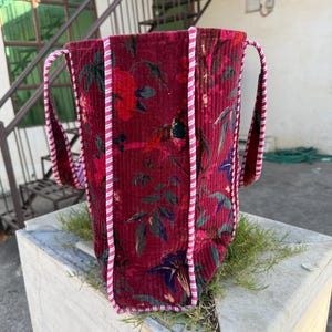 May include: A burgundy corduroy tote bag with a floral pattern in shades of red, pink, and blue. The bag has two handles and is trimmed with pink and white striped piping. The bag is standing upright.