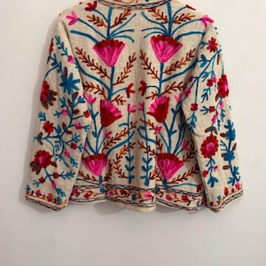 May include: A beige long-sleeved jacket featuring vibrant floral embroidery in shades of hot pink, bright red, and turquoise blue. The design includes symmetrical flower motifs and leafy stems covering the front, back, and sleeves of the garment.
