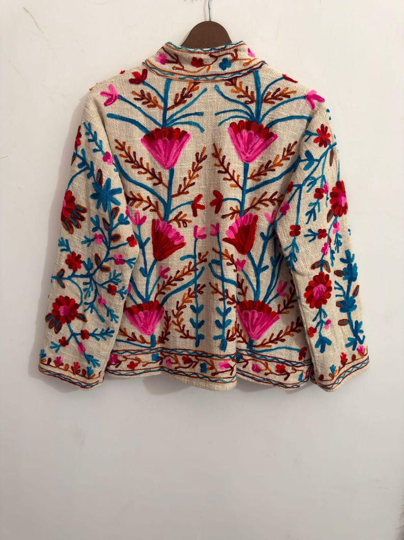 May include: A beige long-sleeved jacket featuring vibrant floral embroidery in shades of hot pink, bright red, and turquoise blue. The design includes symmetrical flower motifs and leafy stems covering the front, back, and sleeves of the garment.