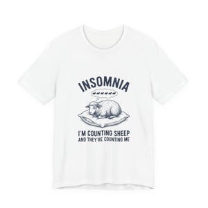 May include: A white short-sleeve t-shirt featuring a dark blue graphic of a sheep lying on a pillow. Above the sheep is the text "INSOMNIA" and a thought bubble with five small sheep. Below is the text "I'M COUNTING SHEEP AND THEY'RE COUNTING ME".