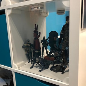 Może przedstawiać: Biała gablota z różnymi figurkami. Figurki przedstawiają postacie w mundurach wojskowych i futurystycznych zbrojach. Gablota ma przezroczysty szklany front i niebieskie panele, eksponujące szczegółowe figurki.