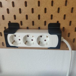 Könnte beinhalten: Ein weißes Mehrfachsteckdosenleiste mit drei Steckdosen und einer schwarzen Halterung ist an einer Lochwand befestigt. Das Mehrfachsteckdosenleiste hat ein weißes Kabel. Die Lochwand ist aus Holz.