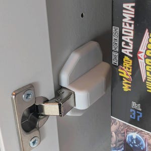 Puede incluir: Un tope de puerta de armario blanco impreso en 3D instalado en una puerta de armario gris. El tope está posicionado cerca de una bisagra plateada y está diseñado para amortiguar el cierre de la puerta. Se ve un volumen de "My Hero Academia".
