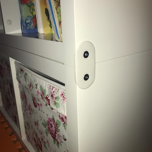 Può includere: Libreria in legno bianco con cerniera in plastica bianca e viti nere. Lo scaffale contiene libri e contenitori in tessuto con un motivo floreale rosa e bianco.