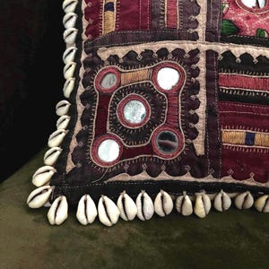 May include: Close-up of a decorative pillow with a patchwork design in burgundy, black, and beige. The pillow features embroidered details, mirrored accents, and is trimmed with white seashells.