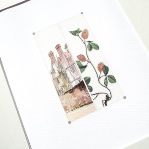 May include: A framed art print featuring a watercolor illustration. The artwork depicts a building with red brick and chimneys, alongside a botanical illustration of a plant with green leaves and reddish-brown seed heads.