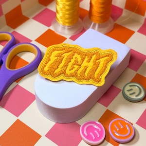 Peut inclure: Un écusson brodé jaune avec le mot "FIGHT" posé sur une forme en mousse blanche. L'image présente également des bobines de fil jaune, des ciseaux violets et des boutons smiley colorés sur un fond à carreaux.