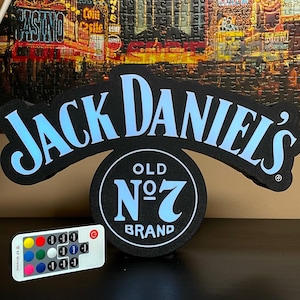 Könnte beinhalten: Ein schwarzes Jack Daniel's Old No. 7 Brand Schild mit hellblauer Schrift und einer passenden Fernbedienung. Das Schild zeigt das ikonische Logo in einer geschwungenen Bogenform über einem kreisförmigen Emblem. Die Fernbedienung enthält Tasten.