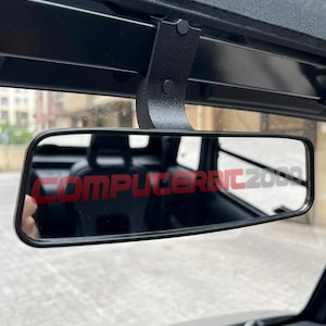 May include: A black rectangular rearview mirror mounted on a black bracket. The mirror features the red text "COMPUTERENC2000" printed across the center of the glass surface, reflecting the interior of a vehicle with a dark seat and headrest.