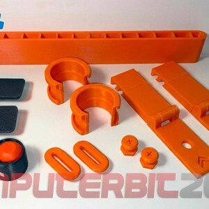 Pode incluir: Um conjunto de componentes de plástico laranja e preto impressos em 3D, incluindo uma bandeja retangular longa, dois clipes em forma de C, duas placas retangulares planas, duas dobradiças, dois anéis ovais, duas pequenas tampas de parafuso e uma base circular.