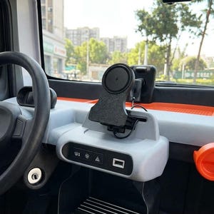 Può includere: Un supporto magnetico circolare nero per telefono fissato al cruscotto grigio chiaro di un veicolo. Il supporto presenta una piccola icona a forma di fulmine. Sotto il supporto si trova un pannello di controllo con icone di sbrinamento.