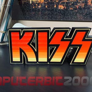 Peut inclure: Une enseigne en trois dimensions affichant le mot KISS dans une police stylisée et audacieuse. Les lettres présentent une bordure noire et un dégradé de couleurs passant du rouge vif en haut à l'orange et au jaune en bas, créant un effet coucher de soleil.