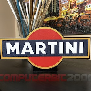 Może przedstawiać: Ozdobny znak z logo Martini, zawierający centralne czerwone koło, poziomy ciemnoniebieski prostokątny pasek oraz słowo MARTINI napisane pogrubionymi białymi wielkimi literami. Projekt posiada cienką złotą obwódkę oraz czarną podstawę do ekspozycji.