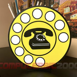 Può includere: Un cartello circolare giallo con una grafica nera di un telefono a disco al centro, circondato da dieci spazi circolari bianchi disposti ad anello. Il testo "COMPUTERBIT 2000" è stampato in un carattere rosso scuro nella parte inferiore.