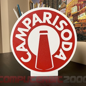 Pode incluir: Uma placa circular com fundo vermelho, borda branca e um círculo branco central. O texto CAMPARISODA está impresso em letras maiúsculas brancas ao redor do centro, que contém uma silhueta vermelha estilizada de uma garrafa.