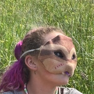 May include: A brown and tan felt mask shaped like a squirrel or chipmunk with star-shaped eye details. The mask has a white elastic band. The person is wearing a beaded necklace with pastel colours.