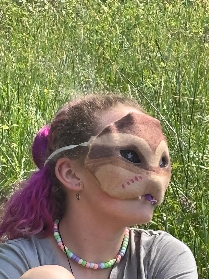 May include: A brown and tan felt mask shaped like a squirrel or chipmunk with star-shaped eye details. The mask has a white elastic band. The person is wearing a beaded necklace with pastel colours.