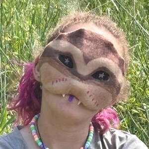 May include: A brown and tan mask with star-shaped eye cutouts and white teeth. The mask is worn by a person with pink hair and a beaded necklace with pastel-coloured beads. The background is a field of green grass.
