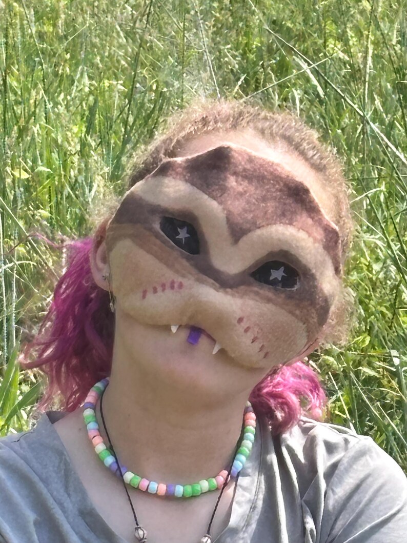 May include: A brown and tan mask with star-shaped eye cutouts and white teeth. The mask is worn by a person with pink hair and a beaded necklace with pastel-coloured beads. The background is a field of green grass.