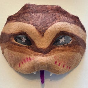 May include: A handcrafted felt mask in brown and beige tones, with dark brown stripes and star-shaped eye details. The mask has a purple tongue, white teeth, and pink accents, forming a whimsical creature design.