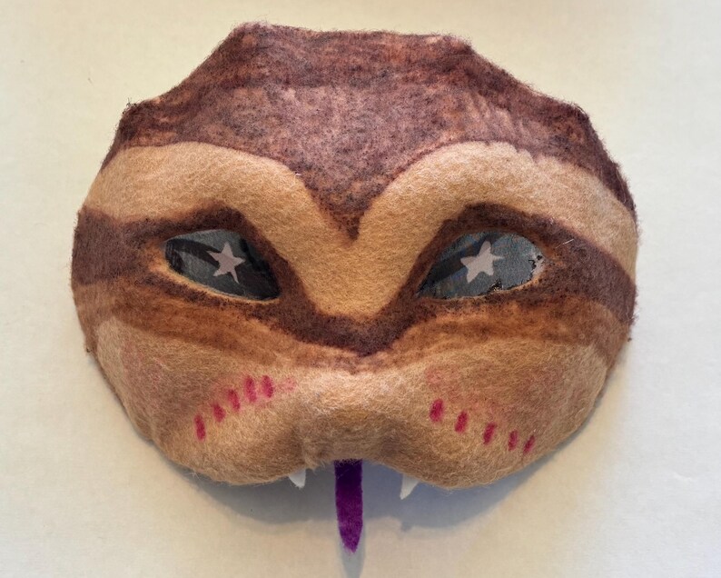 May include: A handcrafted felt mask in brown and beige tones, with dark brown stripes and star-shaped eye details. The mask has a purple tongue, white teeth, and pink accents, forming a whimsical creature design.
