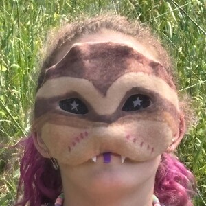 May include: A brown and tan mask with star-shaped eyes and small white teeth. The mask has a purple piece in the mouth area and is worn by a person with pink hair and a colourful beaded necklace. The background is green grass.