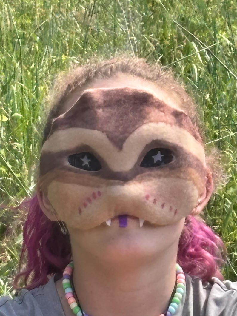 May include: A brown and tan mask with star-shaped eyes and small white teeth. The mask has a purple piece in the mouth area and is worn by a person with pink hair and a colourful beaded necklace. The background is green grass.