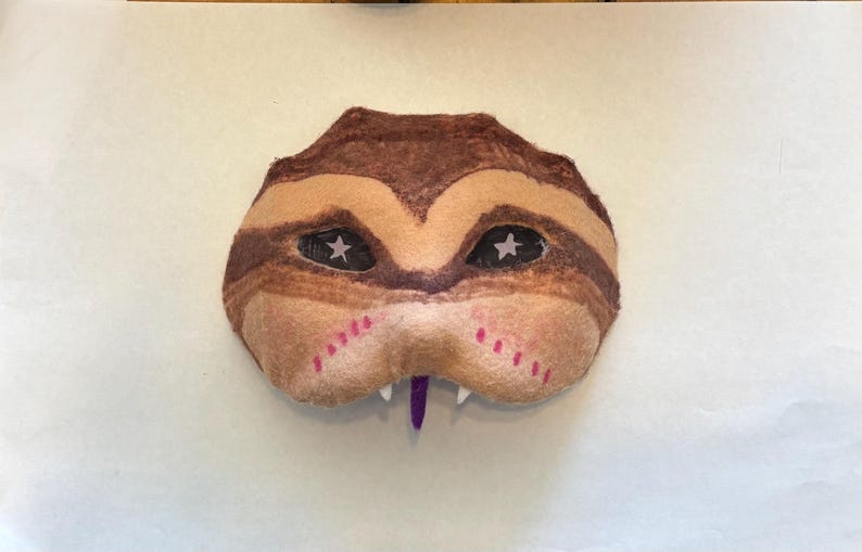 May include: A handmade mask with a reptile-like design. The mask features brown and tan stripes, black eyes with white star accents, and a purple tongue. Pink dotted lines are on the cheeks. The mask is likely for costume or decorative purposes.