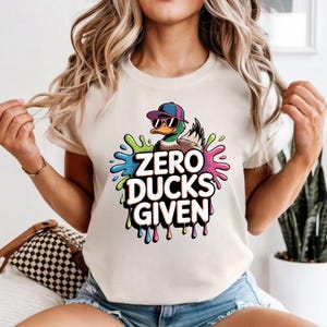May include: Beige t-shirt featuring a cartoon duck wearing sunglasses and a cap, with the text "ZERO DUCKS GIVEN" in a colorful, dripping font. The duck is set against a splash of green, blue, and pink.