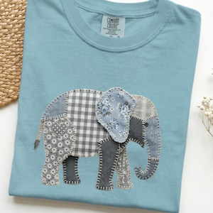 May include: A light blue t-shirt featuring an appliqued elephant design. The elephant is made of various patterned fabrics, including gingham, floral, and solid gray, stitched together. The shirt has a crew neck and short sleeves.