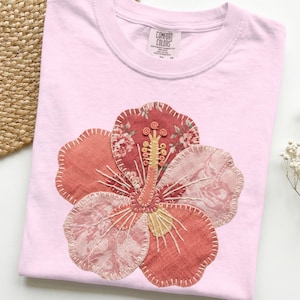 May include: A light pink t-shirt with a large, appliqued hibiscus flower. The flower is made of various fabrics in shades of red, pink, and cream, with detailed stitching. The shirt has a crew neck and a tag that reads "COMFORT COLORS".