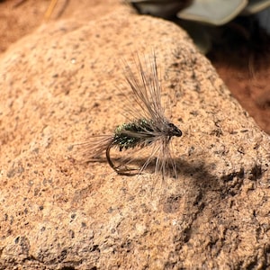 May include: A close-up of a handmade fishing fly. The fly features a dark hook, with a green and brown body, and long, thin, light brown fibers extending outward. The fly is resting on a textured, light brown rock.