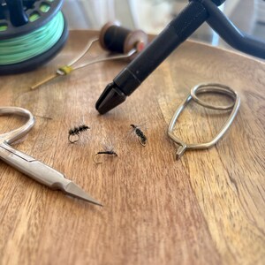 May include: Fly fishing tools and equipment arranged on a wooden surface. Includes a fly reel with green line, a black vise, scissors, thread spools, and several artificial flies. The scene is well-lit, showcasing the details of the angling gear.