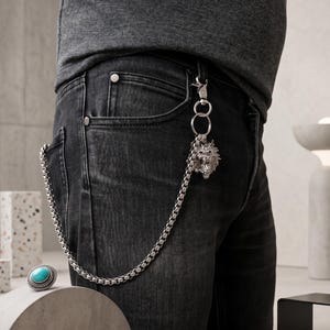 May include: A silver chain wallet chain with a lion head pendant attached to a black belt loop. The chain is connected to a pair of black denim jeans. A turquoise ring is on a white surface.