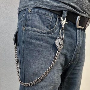 Op de afbeelding: Zilverkleurige portemonnee ketting met een wolfskop hanger. De ketting is gemaakt van dikke, in elkaar grijpende schakels en hangt voor een blauwe spijkerbroek. De riem is zwart met een zilveren gesp.