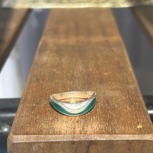 Könnte beinhalten: Ein silberner Ring mit geschwungenem Design und türkisfarbener Emaille-Einlage. Der Ring liegt auf einer Holzoberfläche und unterstreicht seine elegante Schlichtheit. Dieses Schmuckstück ist ein stilvolles Accessoire.