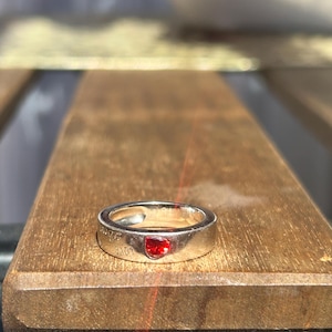Könnte beinhalten: Silberring mit einem roten herzförmigen Stein. Der Ring trägt die Gravur "LOVE". Der Ring liegt auf einer Holzoberfläche.