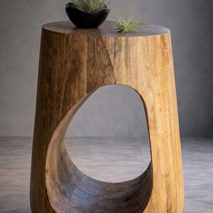 May include: A wooden side table with a distinctive, organic design. The table is made of light brown wood and has a large, curved opening. A small black bowl with greenery sits on top, adding a touch of nature to the decor.