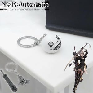 May include: A white, spherical keychain charm with a black, smiling face design. The keychain includes a silver ring and chain. The image also shows a figure of a woman with a sword and the text "NieR:Automata Game of the YoRHa Edition."