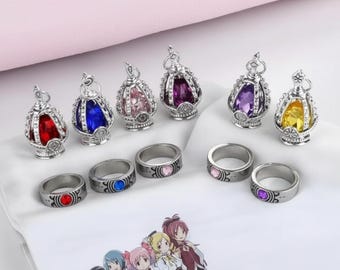 Könnte beinhalten: Eine Sammlung silberfarbener Ringe und Anhänger mit farbigen Edelsteinakzenten. Die Anhänger sind kronenförmig, und die Ringe weisen farblich passende Steine auf. Die Edelsteine sind rot, blau, rosa, lila und gelb.