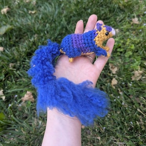 May include: A handmade crochet squirrel with a long, fluffy tail. The squirrel's body is blue and purple, with a yellow head and tiny yellow legs. The tail is a vibrant blue, and the squirrel is held in a hand against a backdrop of green grass.