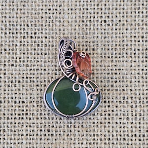 May include: A wire-wrapped pendant featuring a smooth, round, dark green stone shaped like a pumpkin. The copper wire forms intricate swirls and loops around the stone, topped with a small, translucent, amber-colored leaf accent.