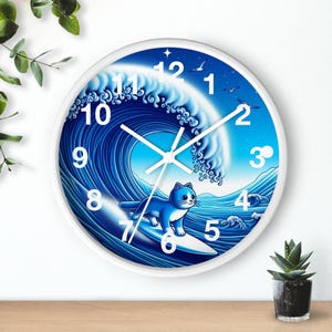 May include: A round wall clock with a white frame featuring a graphic illustration of a blue cat surfing on a large ocean wave. The clock face displays white Arabic numerals from 1 to 12, with white hour, minute, and second hands centered.