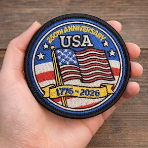 May include: A round patch with a blue border and the text "250TH ANNIVERSARY USA". The patch features a waving American flag and the years "1776-2026" on a yellow banner. The patch is held in a hand.
